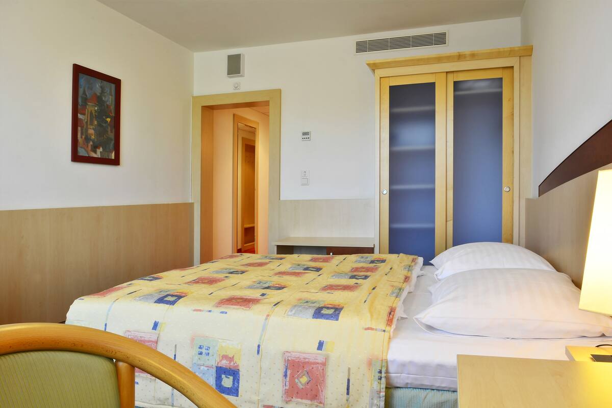 A hotel room with a bed, a chair, a desk, and a painting on the wall.
