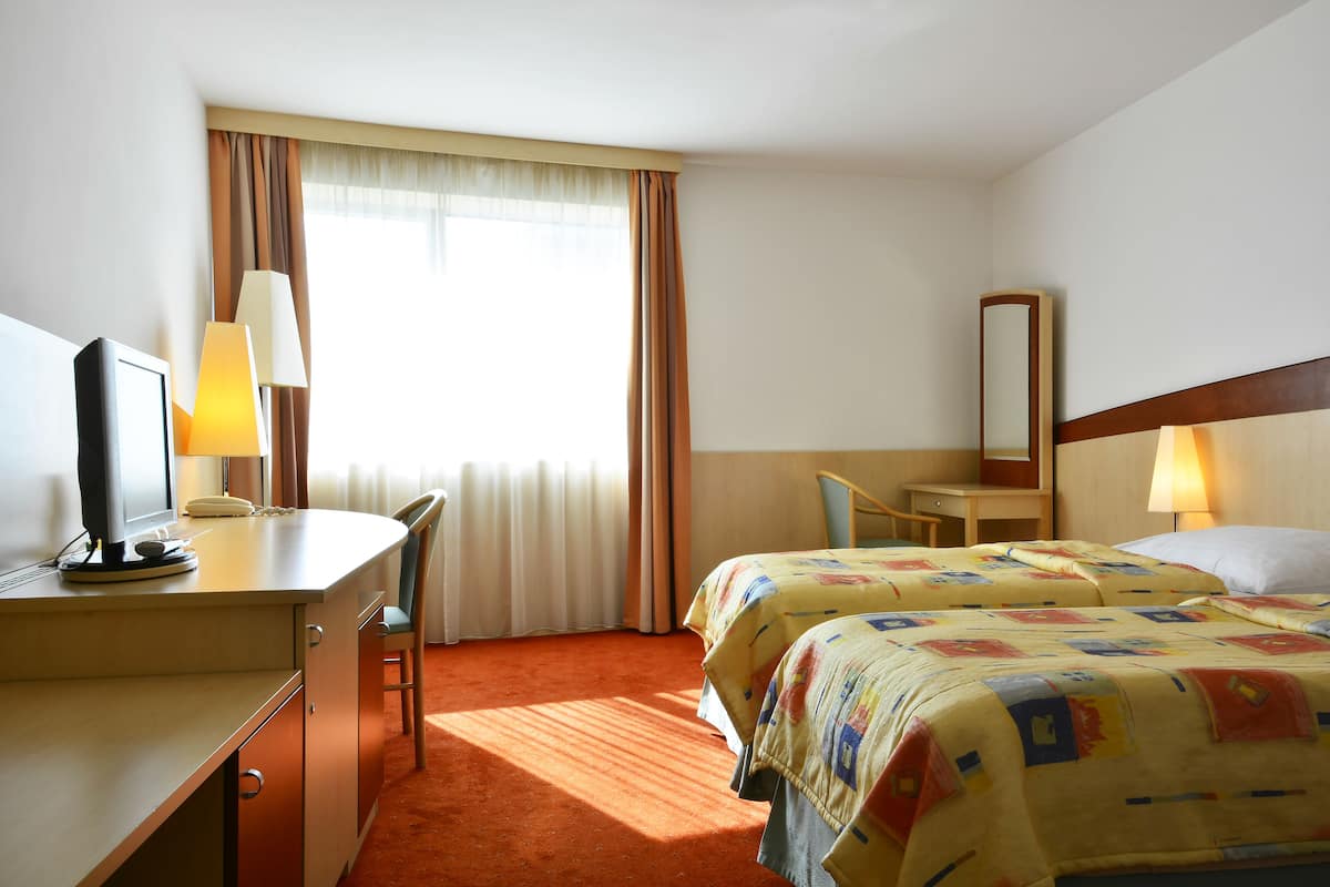 A hotel room with a desk, two beds, a chair, a lamp, and a window with curtains.