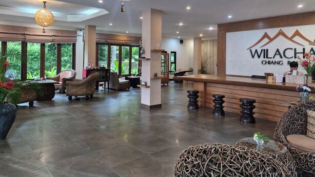 A modern reception area with a reception desk, wicker furniture, and a large window offering a view of greenery. - Wilacha Chiang Rai (Chiang Rai)