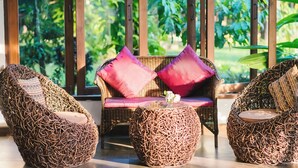 Wicker furniture with cushions and a glass table in a room with large windows and a view of greenery. - Wilacha Chiang Rai (Chiang Rai)