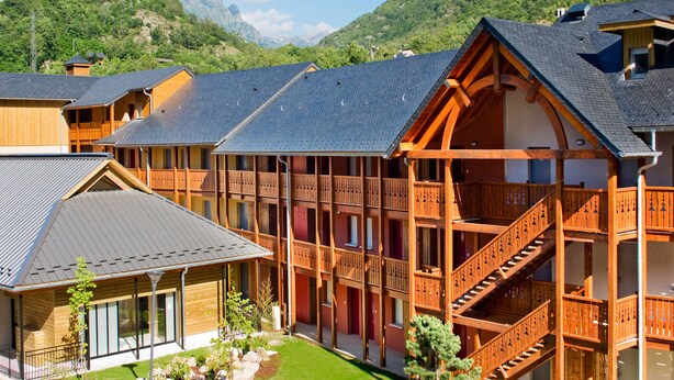 Un bâtiment de style chalet en bois, doté de nombreux balcons et d’un escalier, entouré d’une végétation luxuriante et de montagnes.
