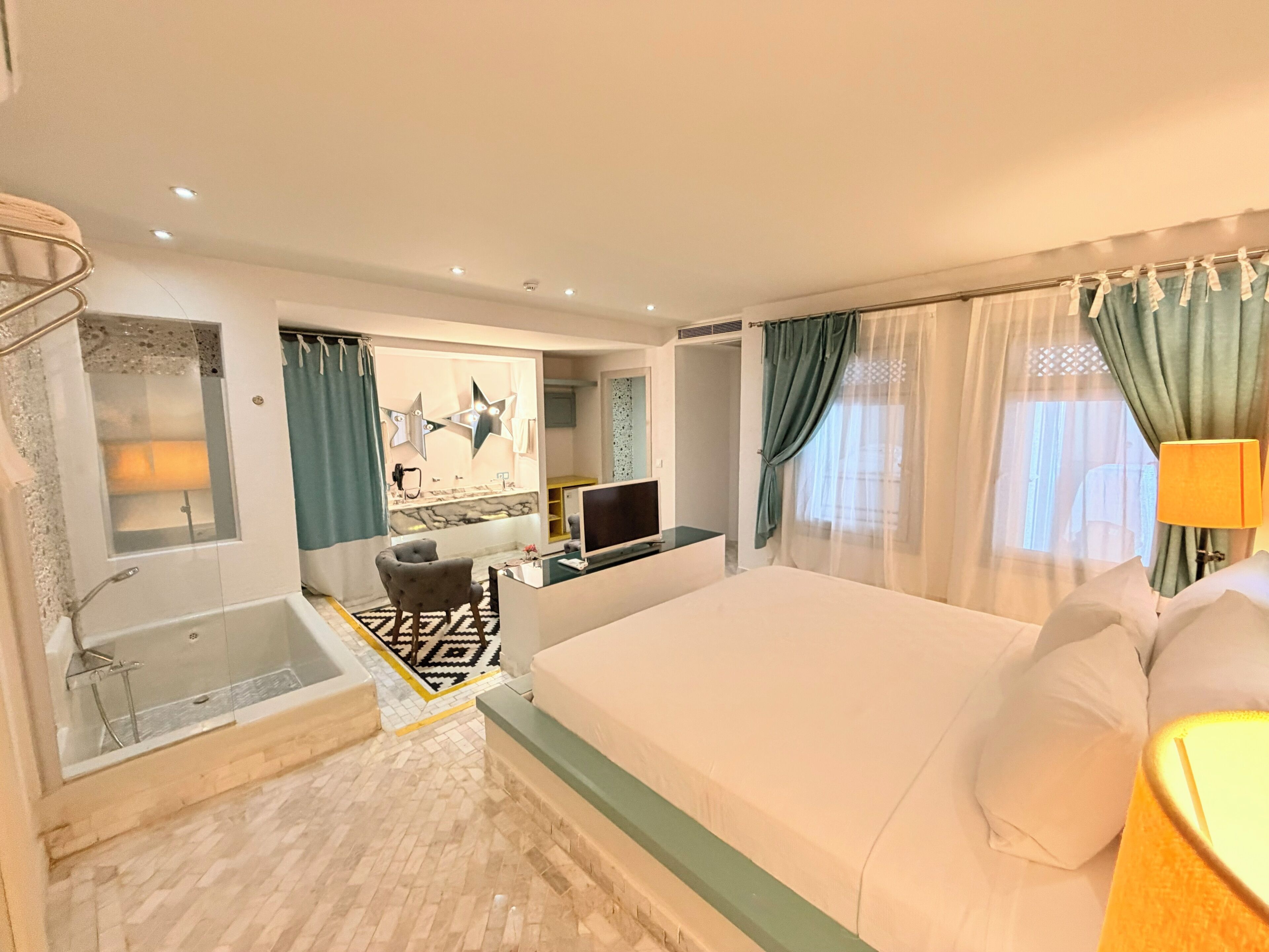 Habitación de hotel con una cama grande, ducha, tocador con espejo, escritorio con televisor y ventana con cortinas.