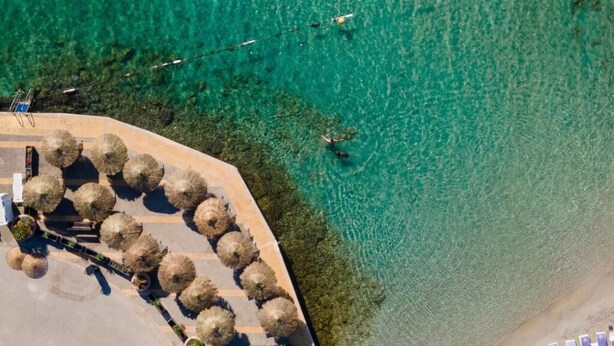 Vista aérea de una zona costera con agua turquesa cristalina, un paseo pavimentado y sombrillas de paja.