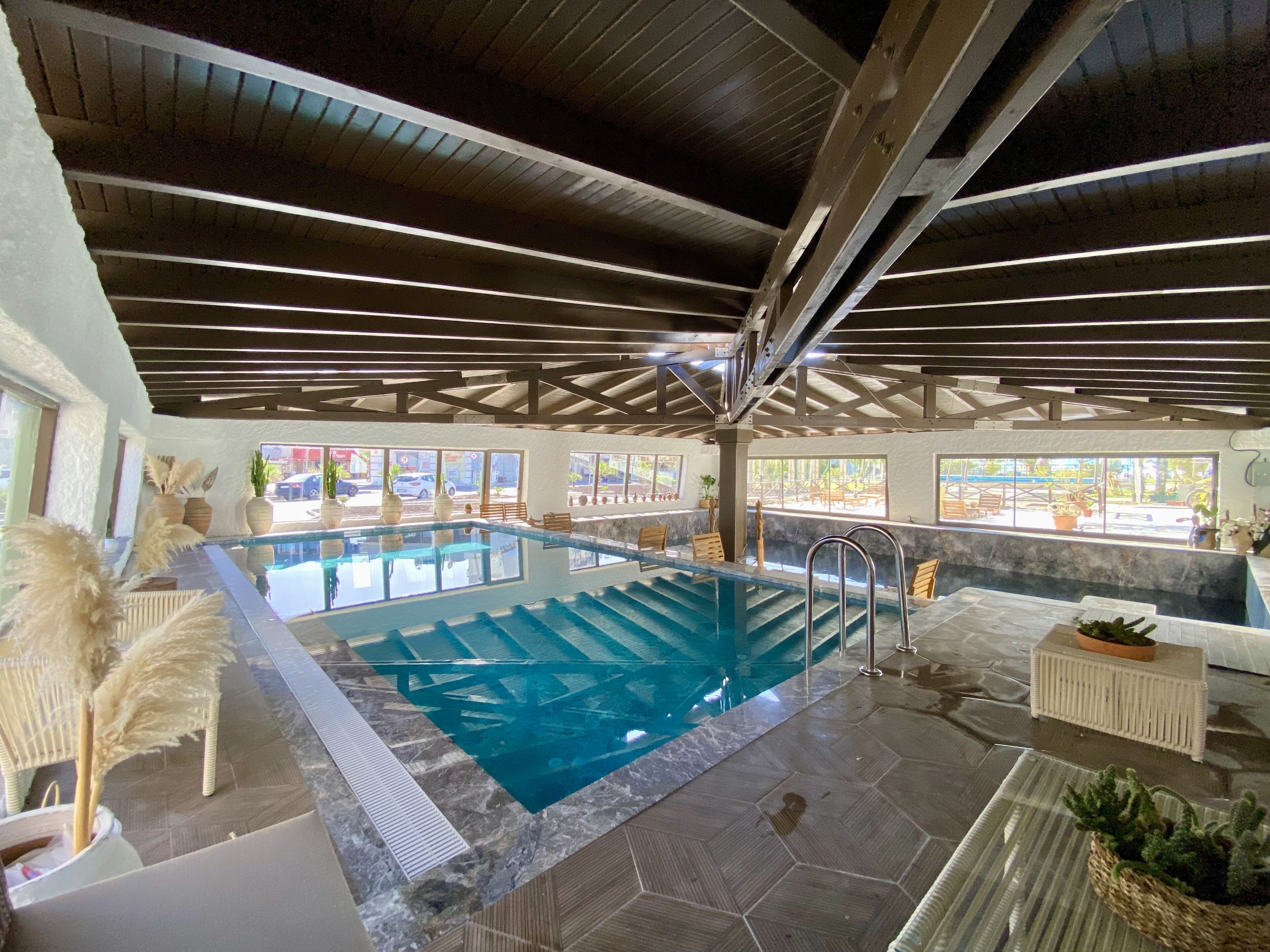 An indoor swimming pool with a view of the outside, surrounded by potted plants and a seating area.