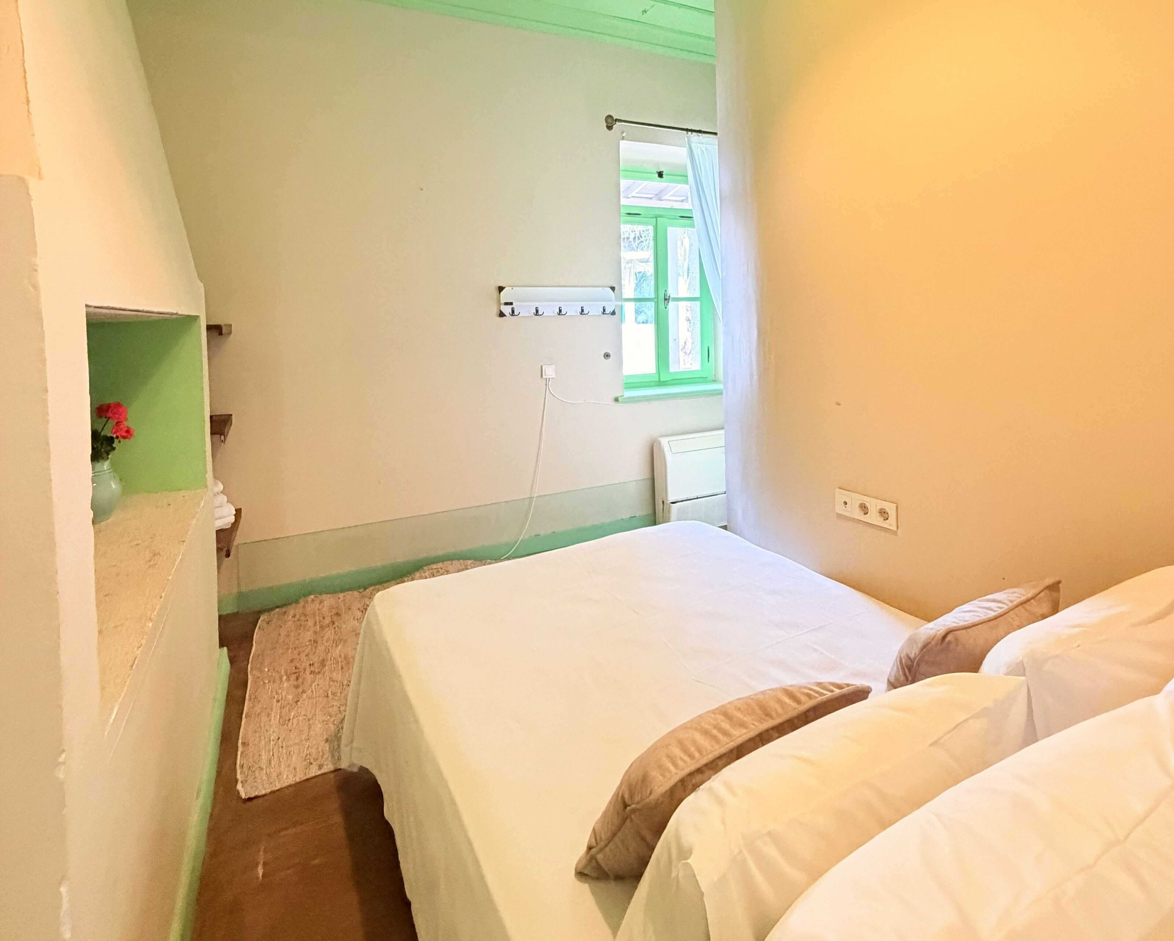 A bedroom with a bed, pillows, a small shelf with flowers, a window with curtains, and a wall-mounted power outlet.