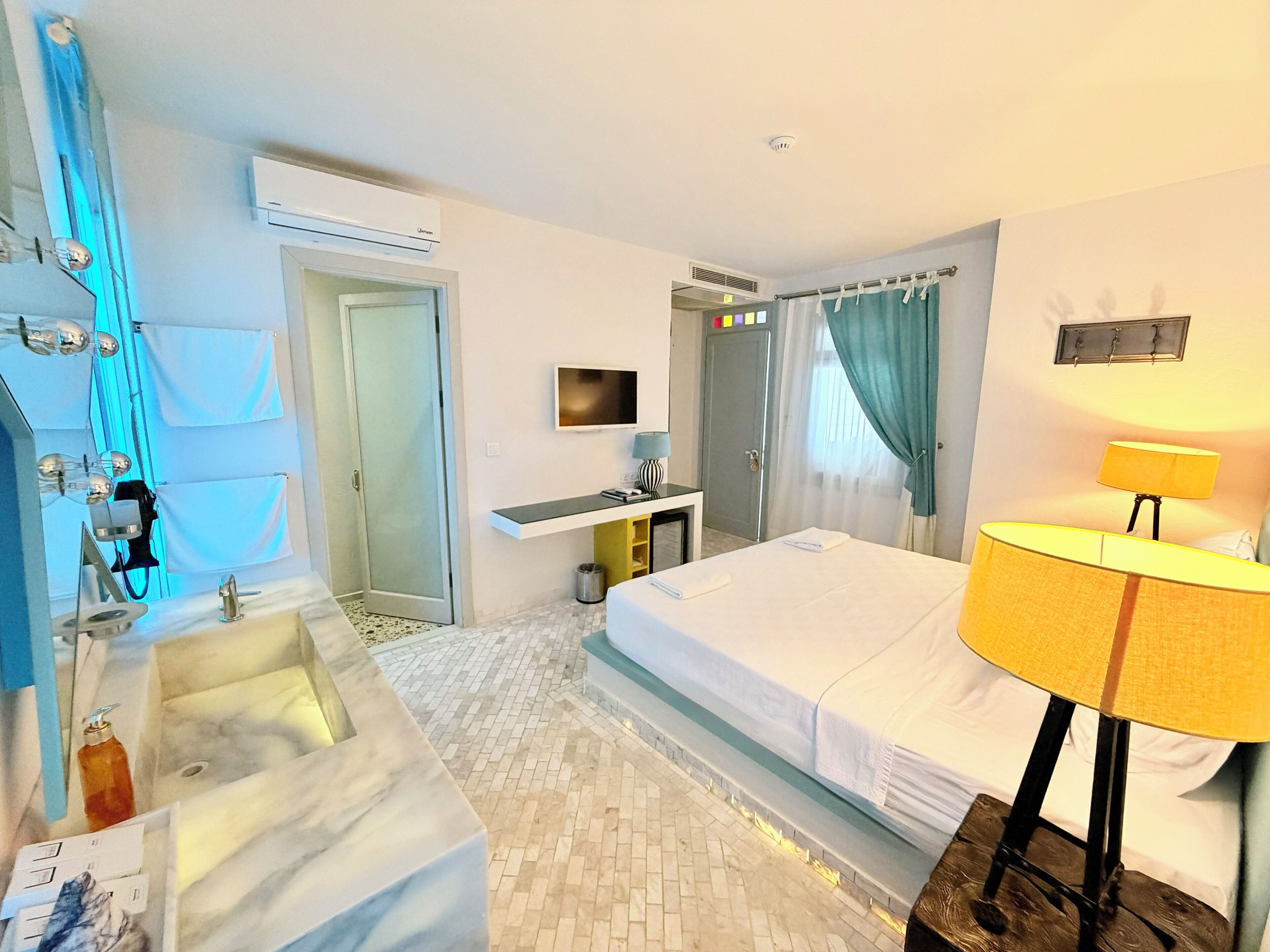 A hotel room with a bed, a desk, a mirror, a TV, and a bathroom with a sink and shower.