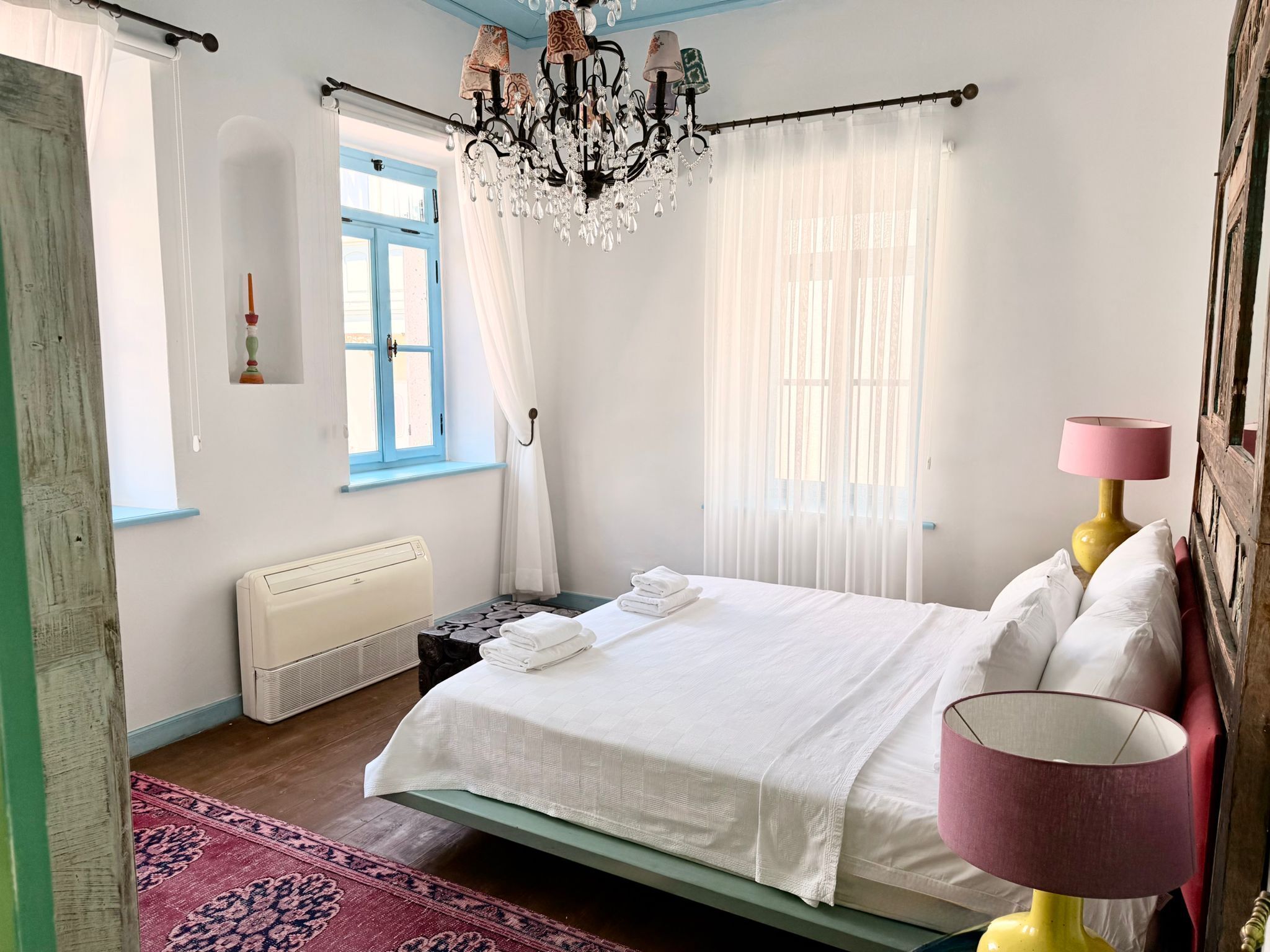 A bedroom with a bed, a chandelier, a window, a radiator, and a lamp.
