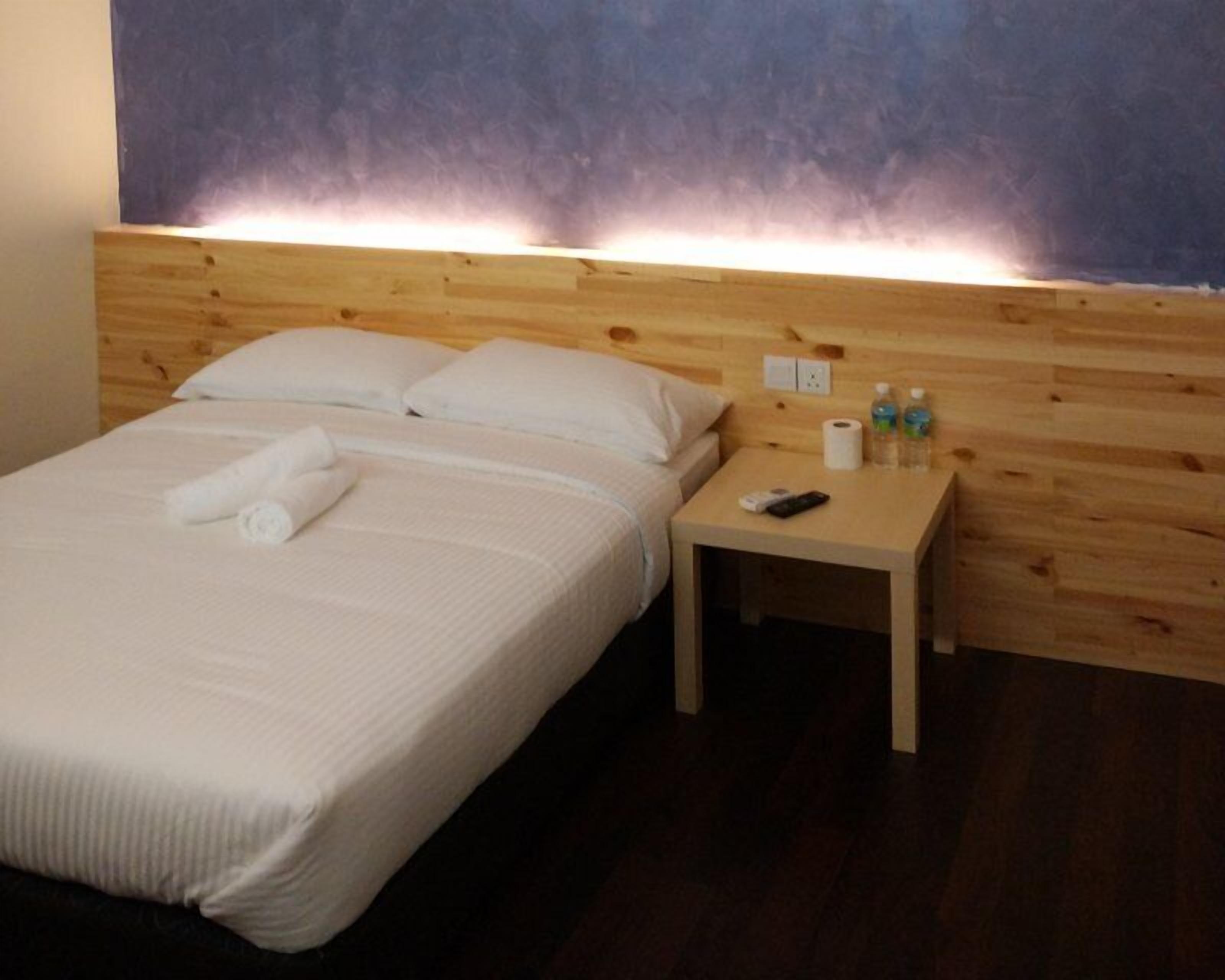 Ein Hotelzimmer mit Holz-Kopfteil, einem ordentlich bezogenen Bett mit weißen Leinen, einem kleinen Nachttisch mit Pflegeartikeln und einer schlichten Wand.