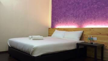 A neatly made bed with white linens and rolled towels, a wooden headboard, and a nightstand with two bottles and a remote control.