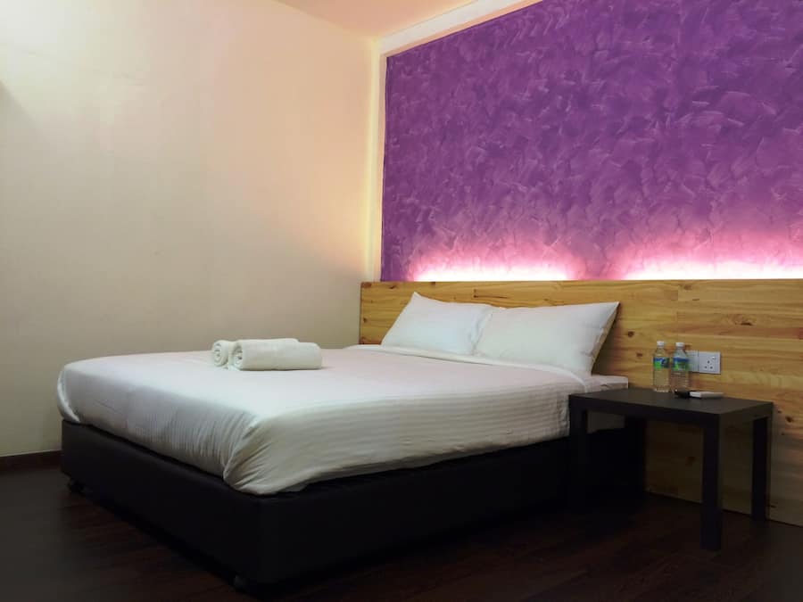 A neatly made bed with white linens and rolled towels, a wooden headboard, and a nightstand with two bottles and a remote control.