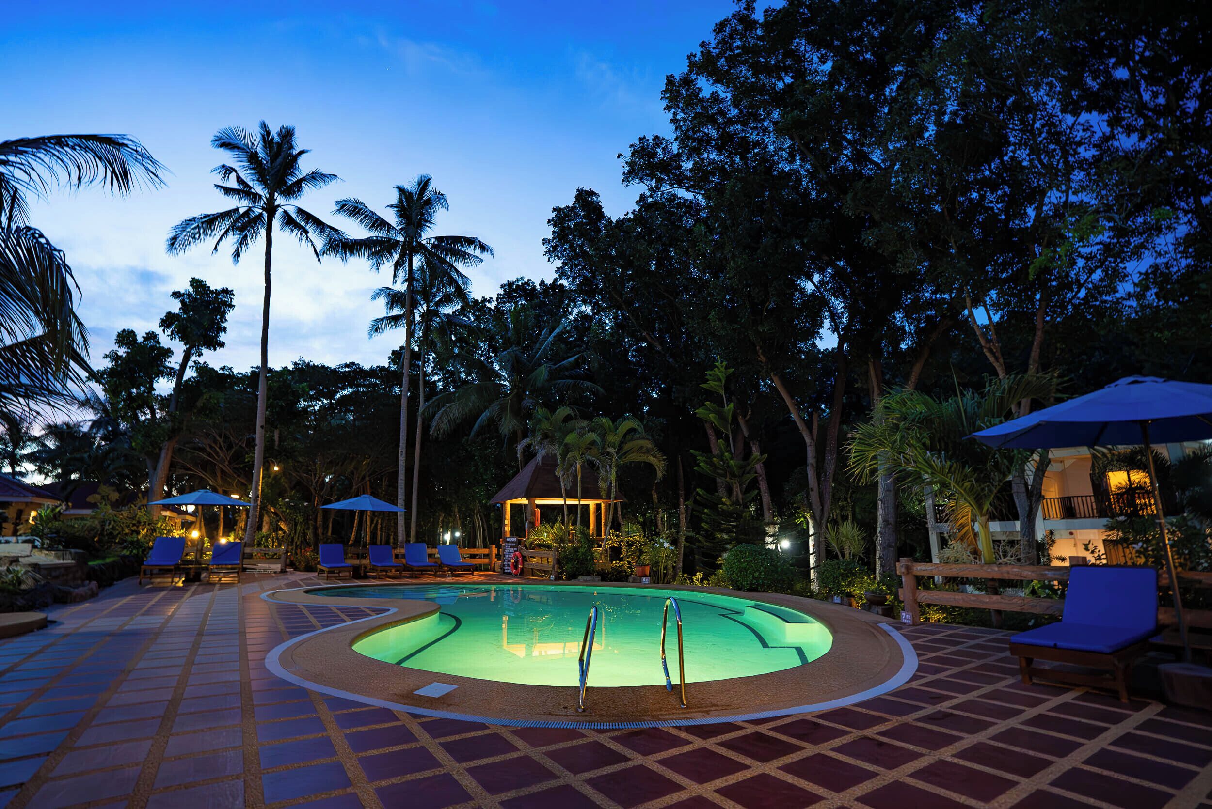 A pool area with lounge chairs and umbrellas, surrounded by palm trees and a gazebo.