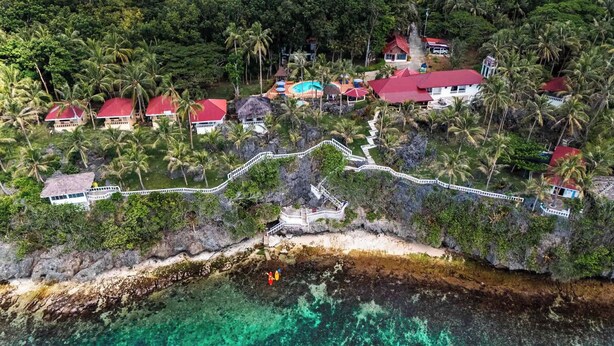 An aerial view of a coastal resort with multiple buildings, a white-walled pathway, and clear turquoise waters. - Blue Star Dive & Resort (Anda)