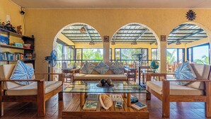 A spacious lounge area with a glass coffee table, two sofas with patterned cushions, and a bookshelf filled with books. - Blue Star Dive & Resort (Anda)