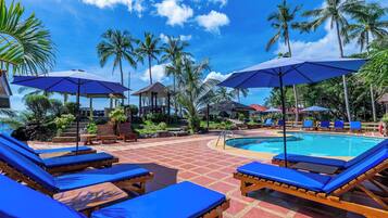A poolside area with blue lounge chairs and umbrellas, surrounded by palm trees and a clear sky.
