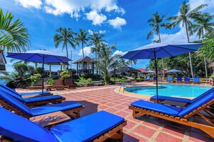 A poolside area with blue lounge chairs and umbrellas, surrounded by palm trees and a clear sky.