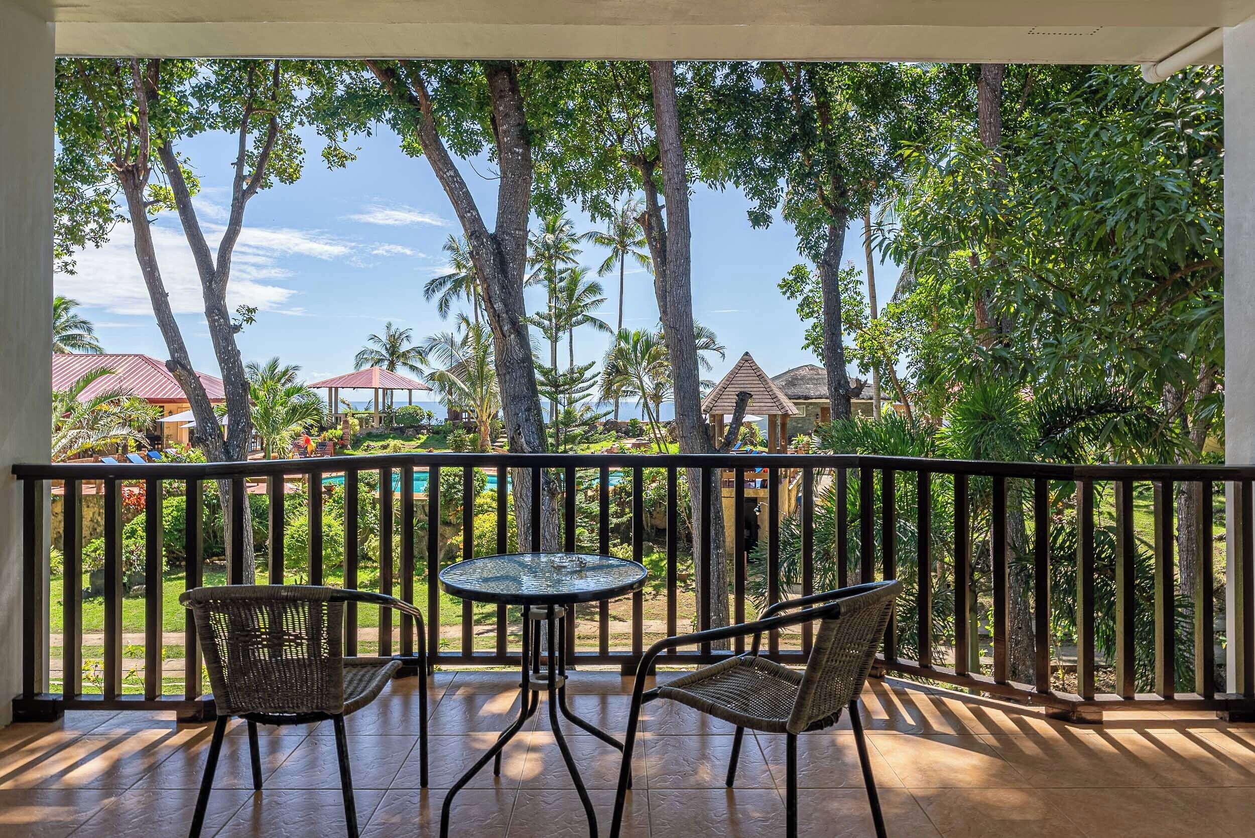 A balcony with two wicker chairs and a small round table, overlooking a tropical garden and a pool area.