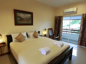 A hotel room with a large bed, a painting on the wall, an air conditioning unit, a window with curtains, and a nightstand with a lamp.