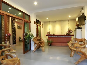 A reception area with a wooden reception desk, two wooden chairs, a potted plant, and a wall-mounted television.