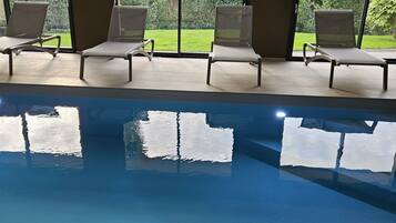 An indoor swimming pool with four lounge chairs and large windows offering a view of greenery.