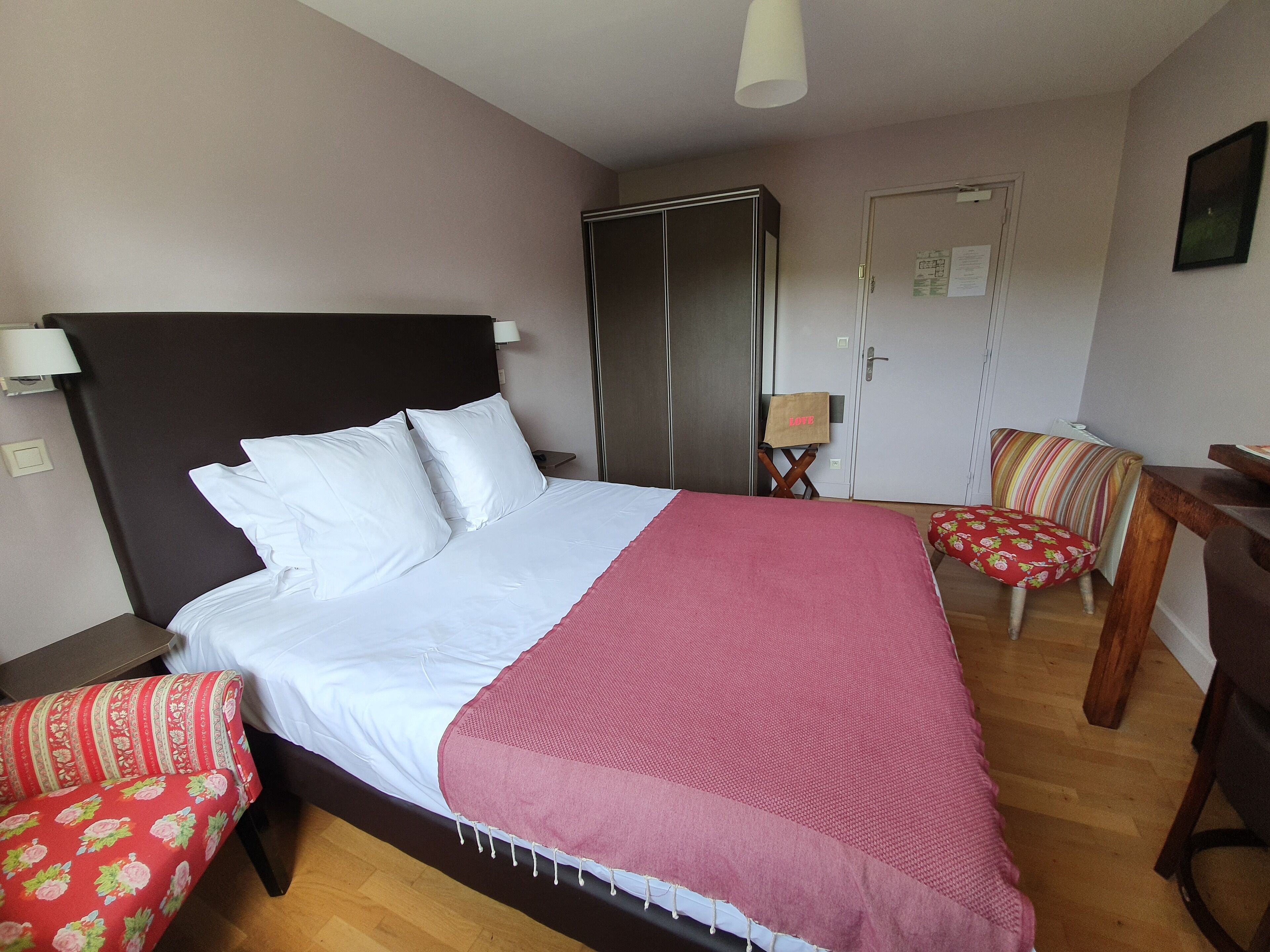 Een net opgemaakt bed met witte lakens en een roze deken, een houten nachtkastje met een lamp, een rode bloemenstoel, een houten bureau en een garderobe.