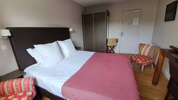 Een net opgemaakt bed met witte lakens en een roze deken, een houten nachtkastje met een lamp, een rode bloemenstoel, een houten bureau en een garderobe.