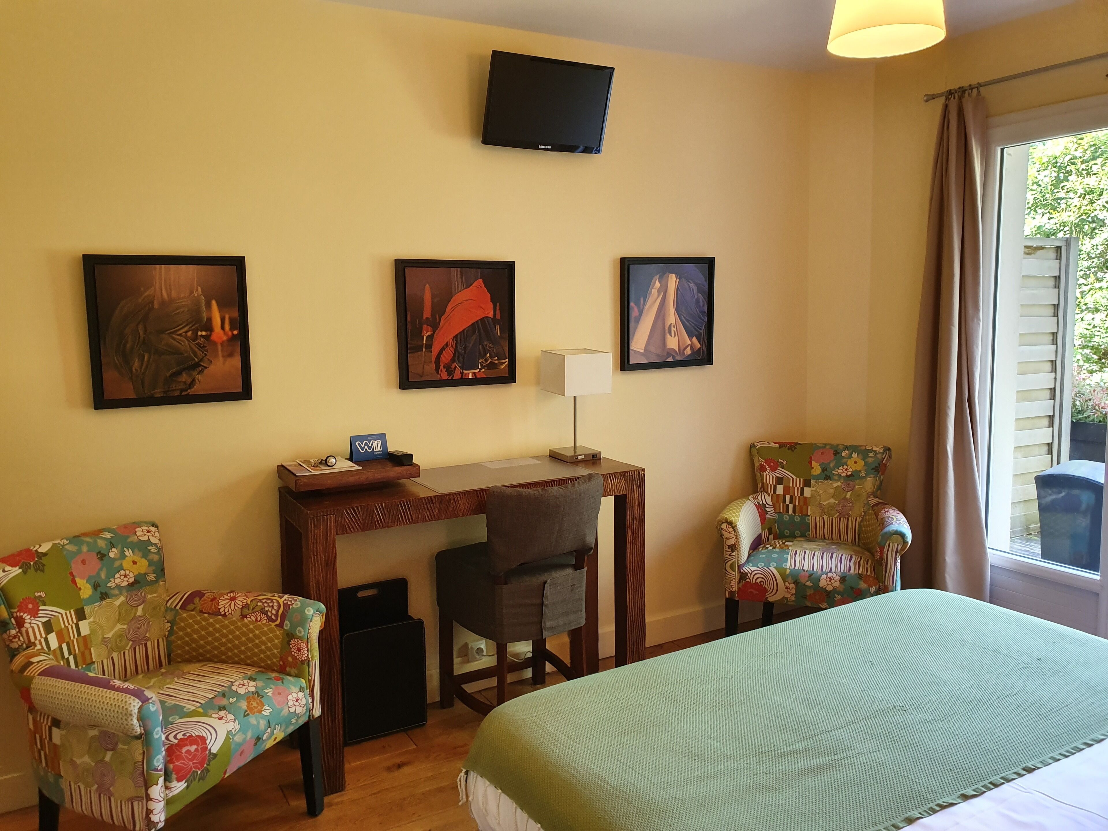 Ein Hotelzimmer mit einem Bett, zwei armchairs mit Blumenmuster, einem Holztisch mit Lampe und drei gerahmten Kunstwerken an der Wand.