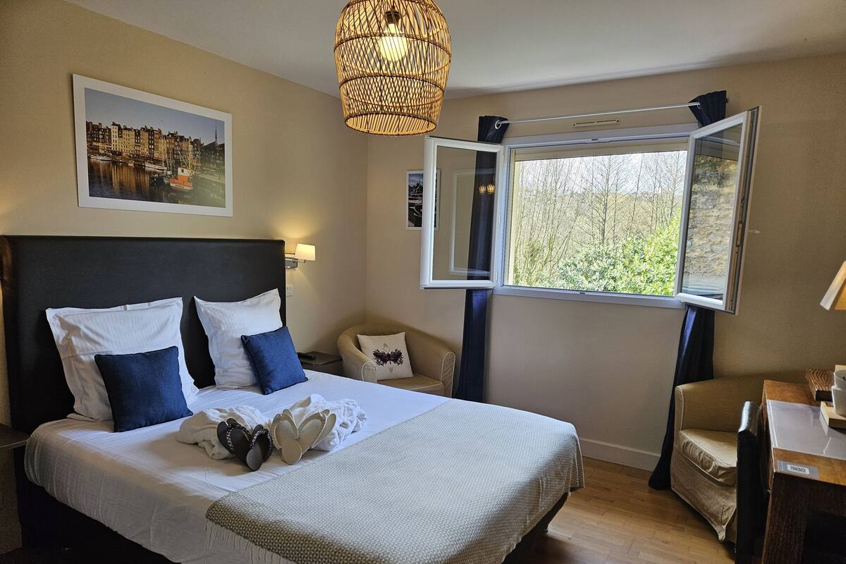 A neatly arranged bedroom with a large bed, a bedside table, a chair, a window with a view of greenery, and a hanging light fixture.