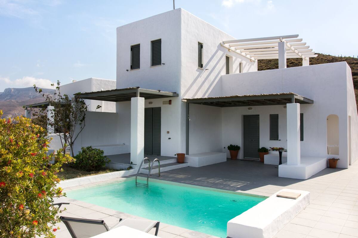 A modern white building with a swimming pool, outdoor seating, and potted plants.