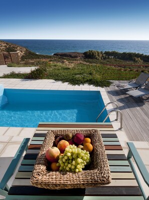 Zona junto a la piscina con una cesta de mimbre llena de fruta, sillones de descanso y vista al mar.