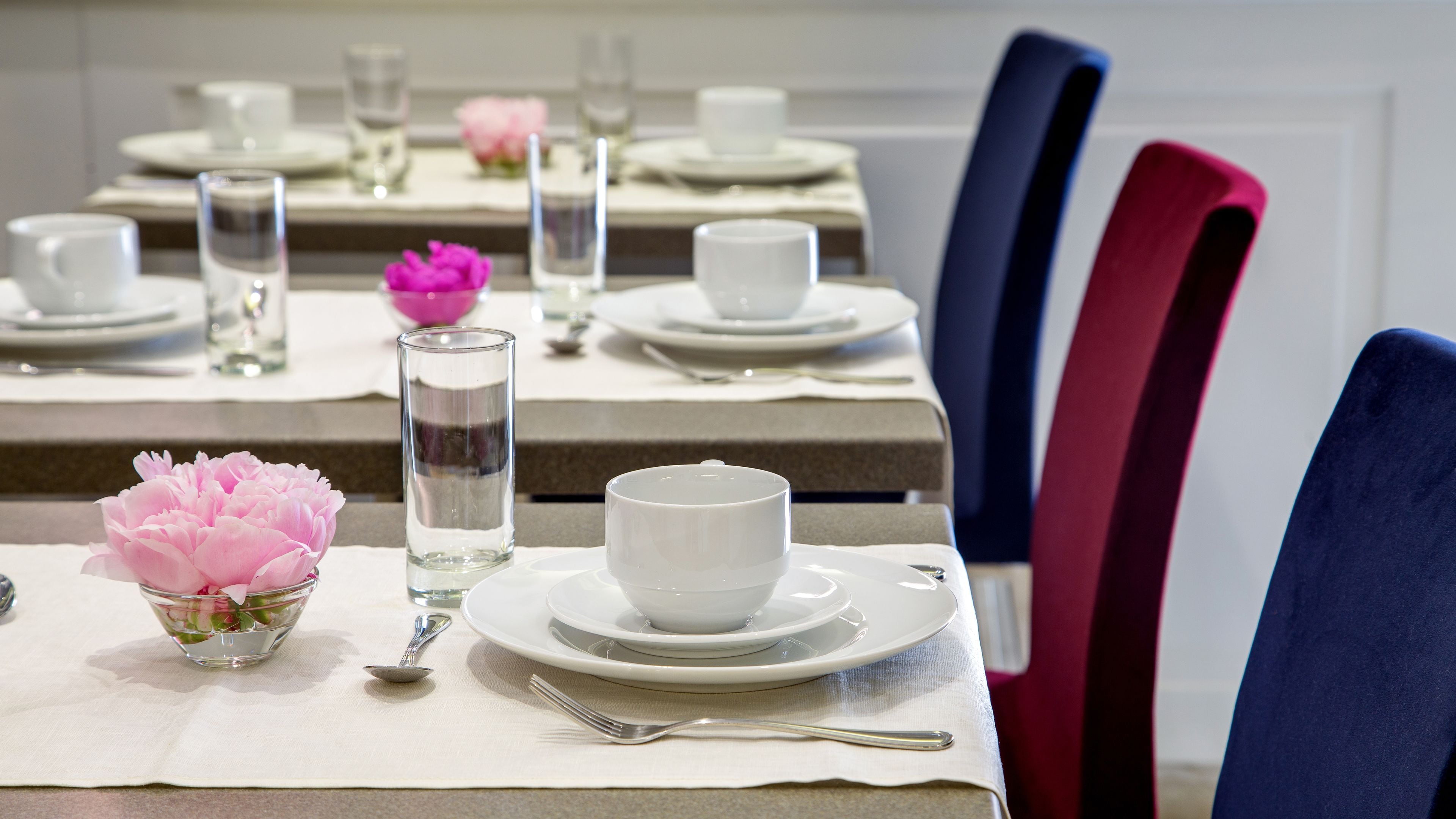 A dining table set with white plates, cups, and a clear glass. There is a pink flower in a glass bowl on the table.