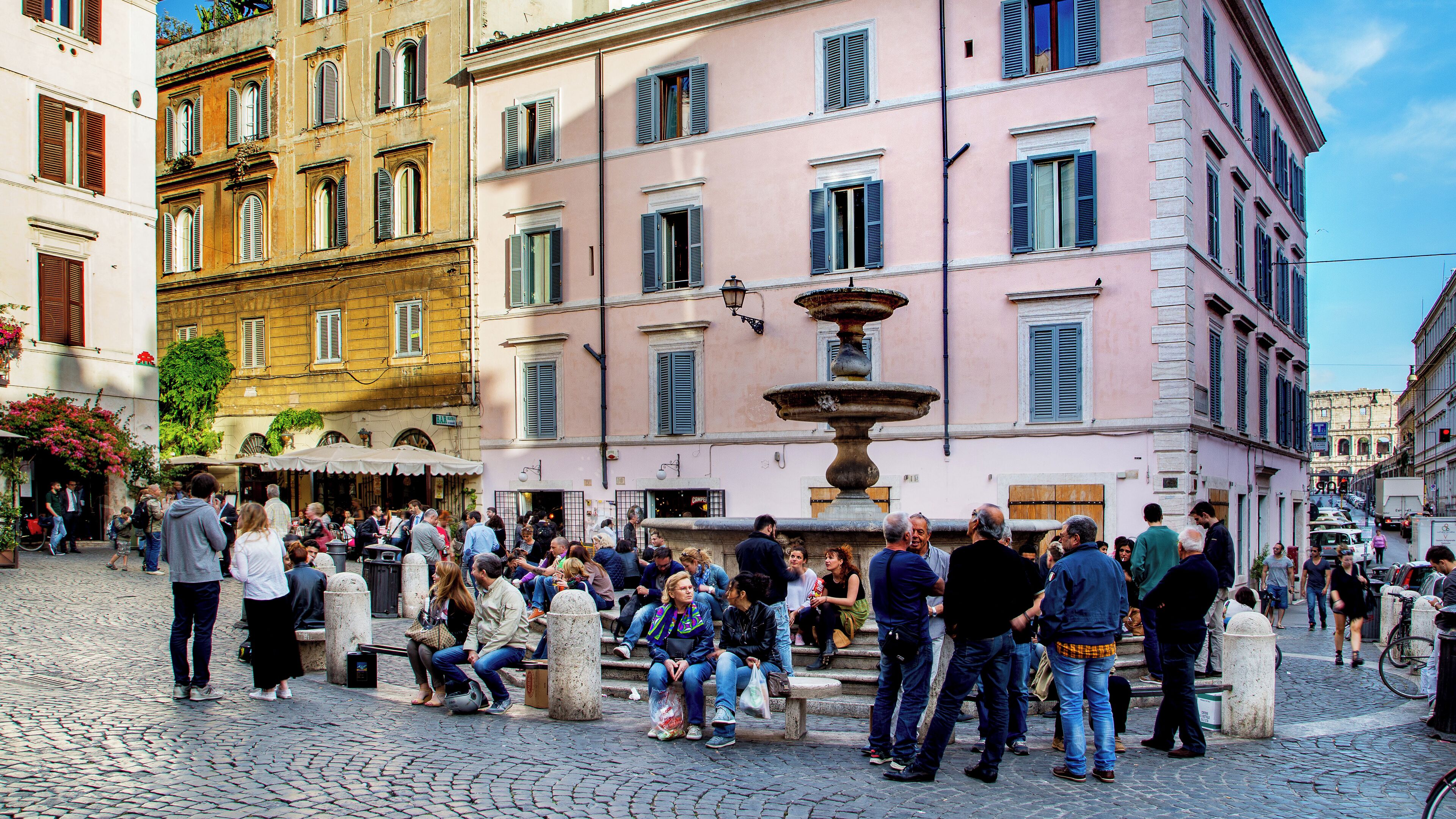 A public square with a fountain, cobblestone pavement, and historic buildings with multiple stories and balconies.