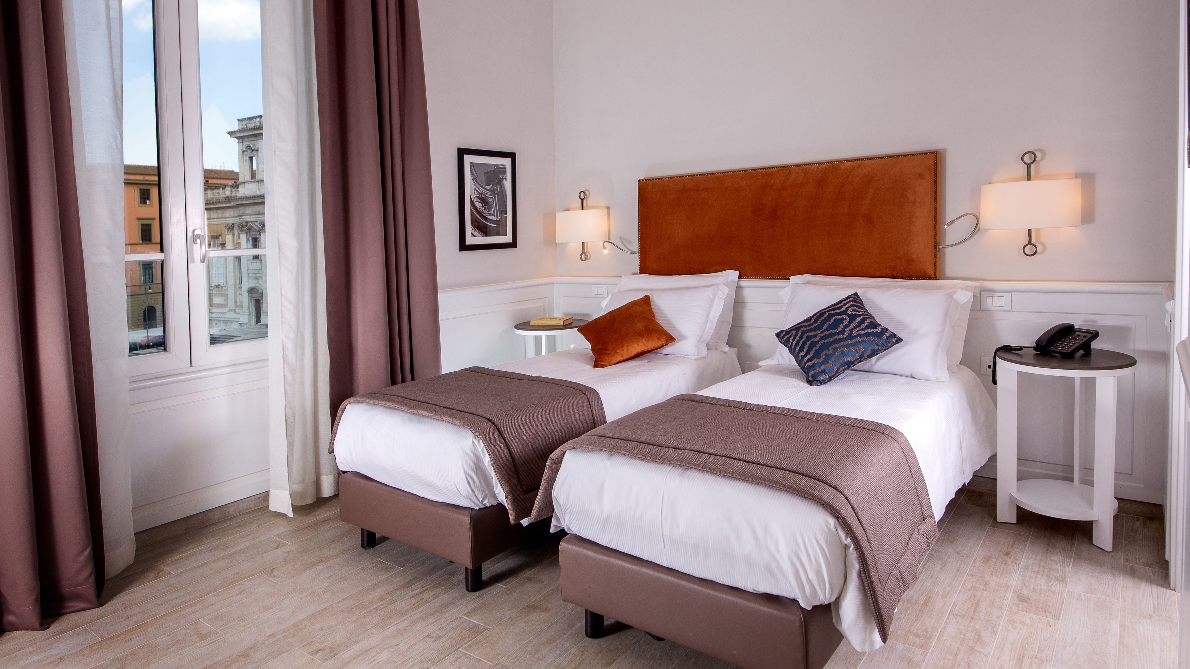 Quarto de hotel com duas camas, uma mesa de cabeceira com abajur, um telefone e vista para a cidade pela janela.