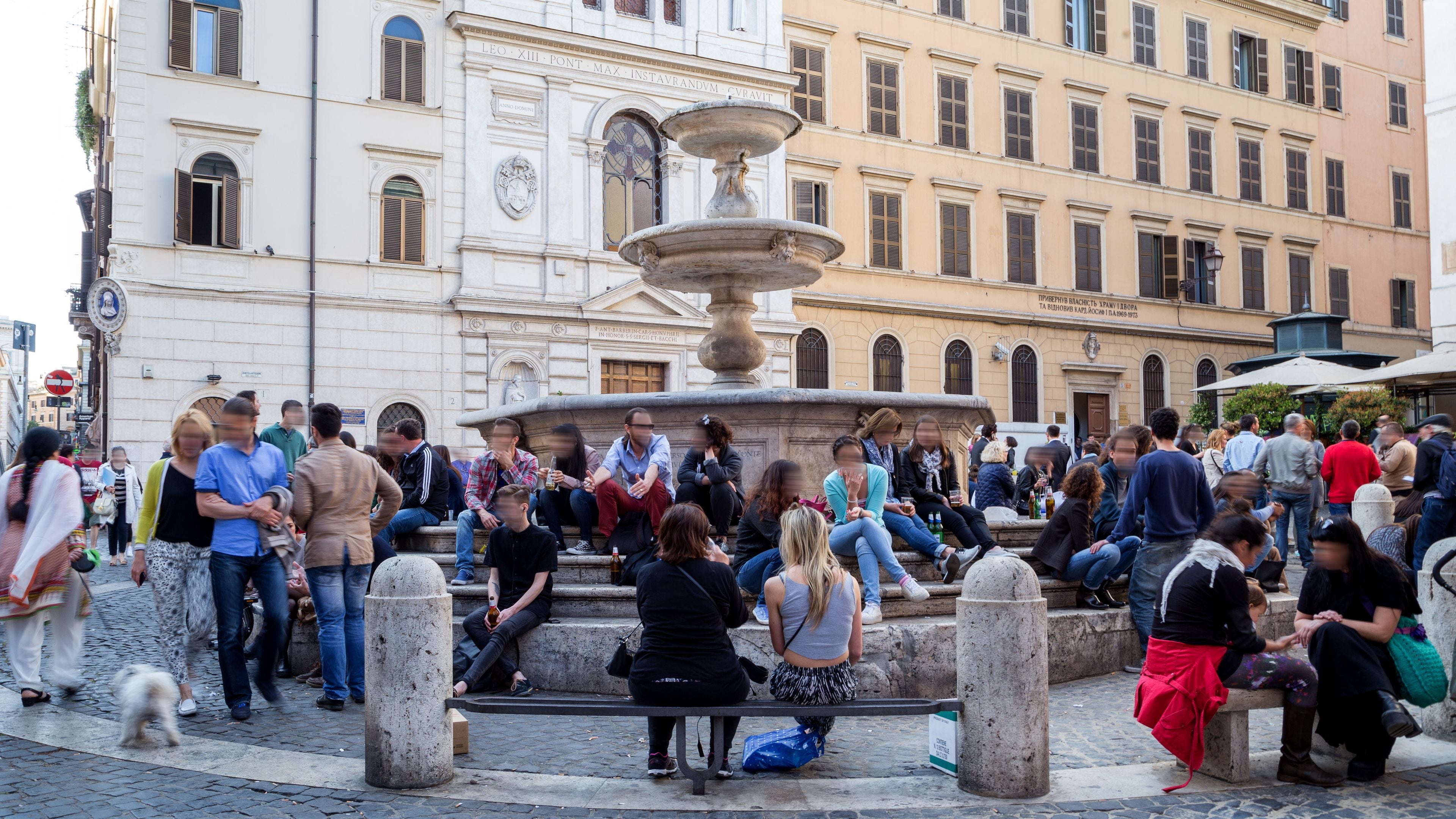 A public square with a central fountain, surrounded by people sitting and standing.