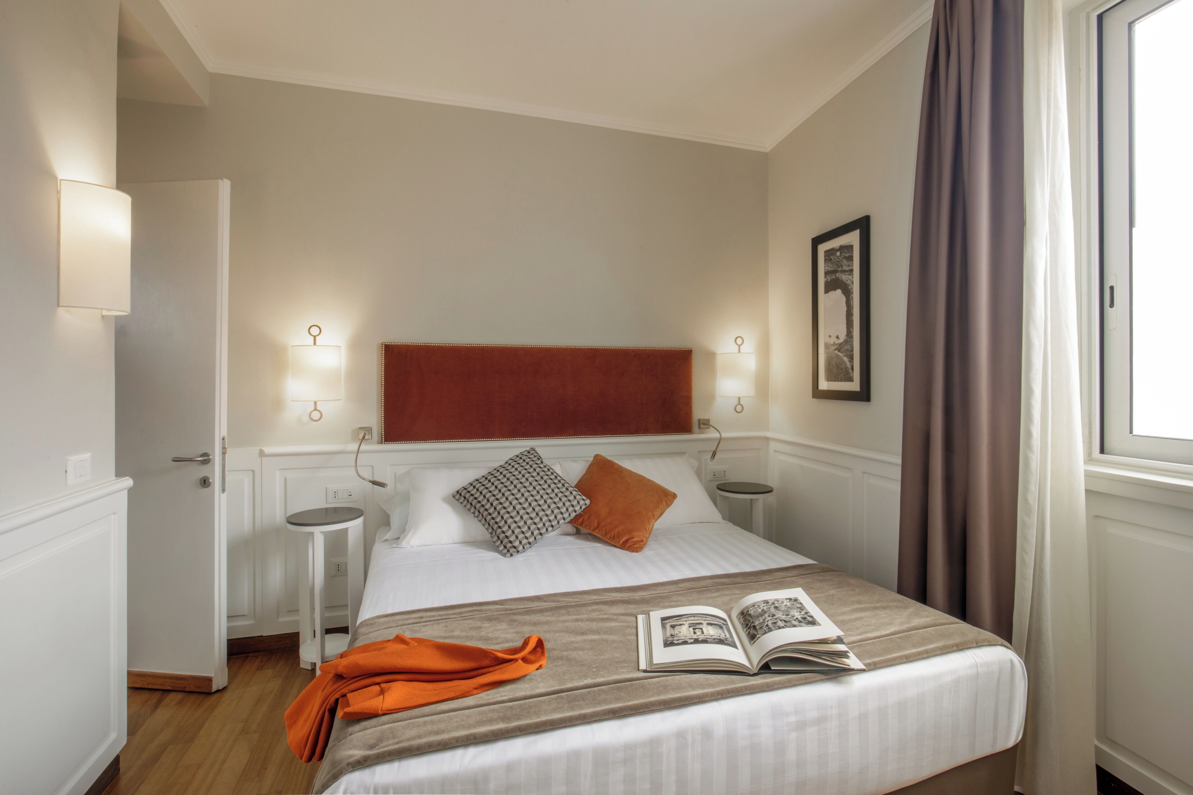 A neatly arranged hotel room with a bed, bedside tables, a reading lamp, a framed picture on the wall, and a window with curtains.