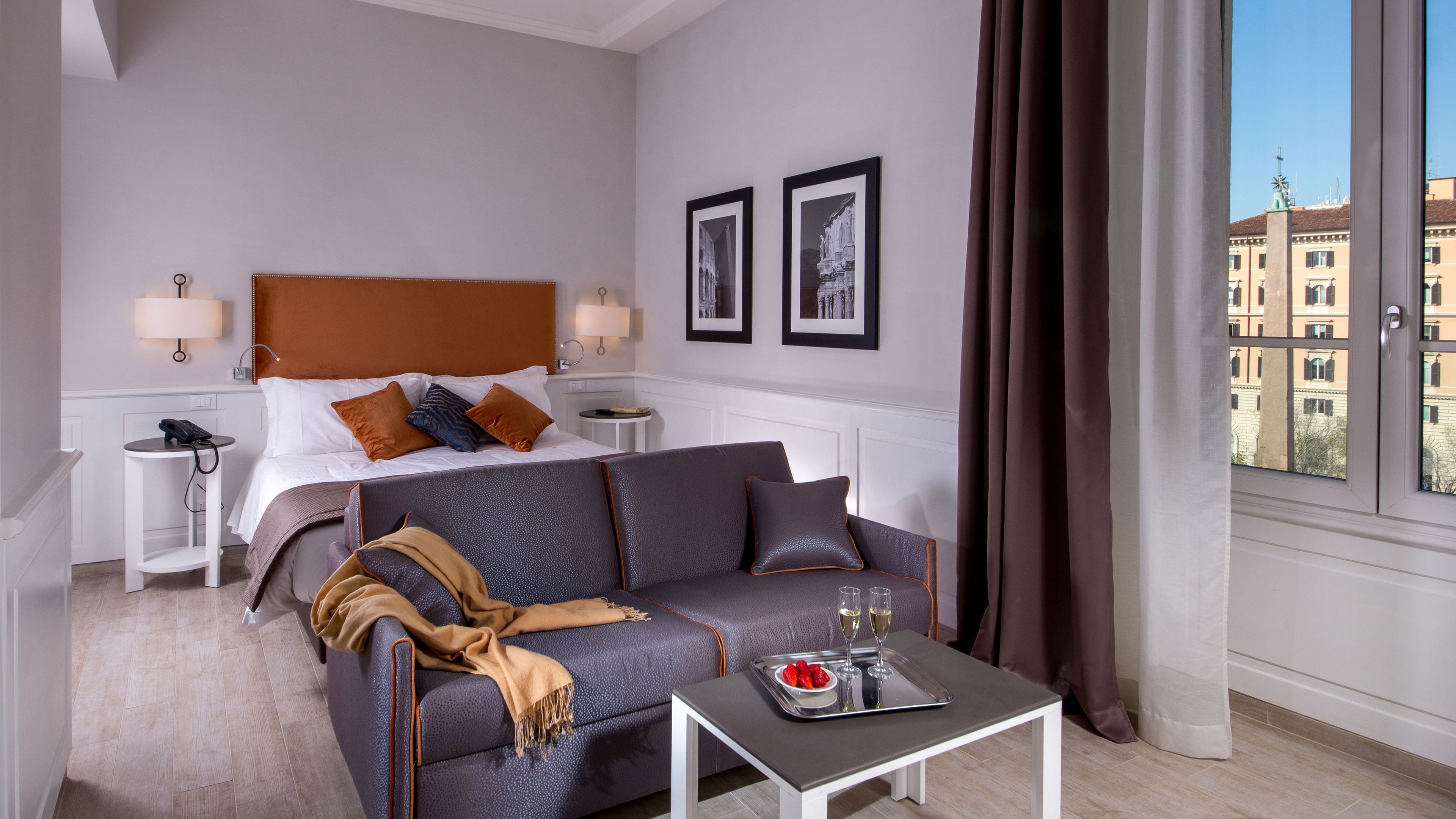 A hotel room with a bed, a gray sofa, a small table with a tray of strawberries and glasses, and a view of buildings through the window.