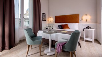 A hotel room with a bed, two chairs, a small table, a bedside table with a phone, and a view of a building through the window.