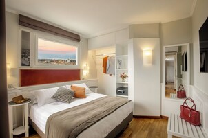 A bedroom with a bed, bedside table, nightstand, wardrobe, mirror, and a view of the city through a window.