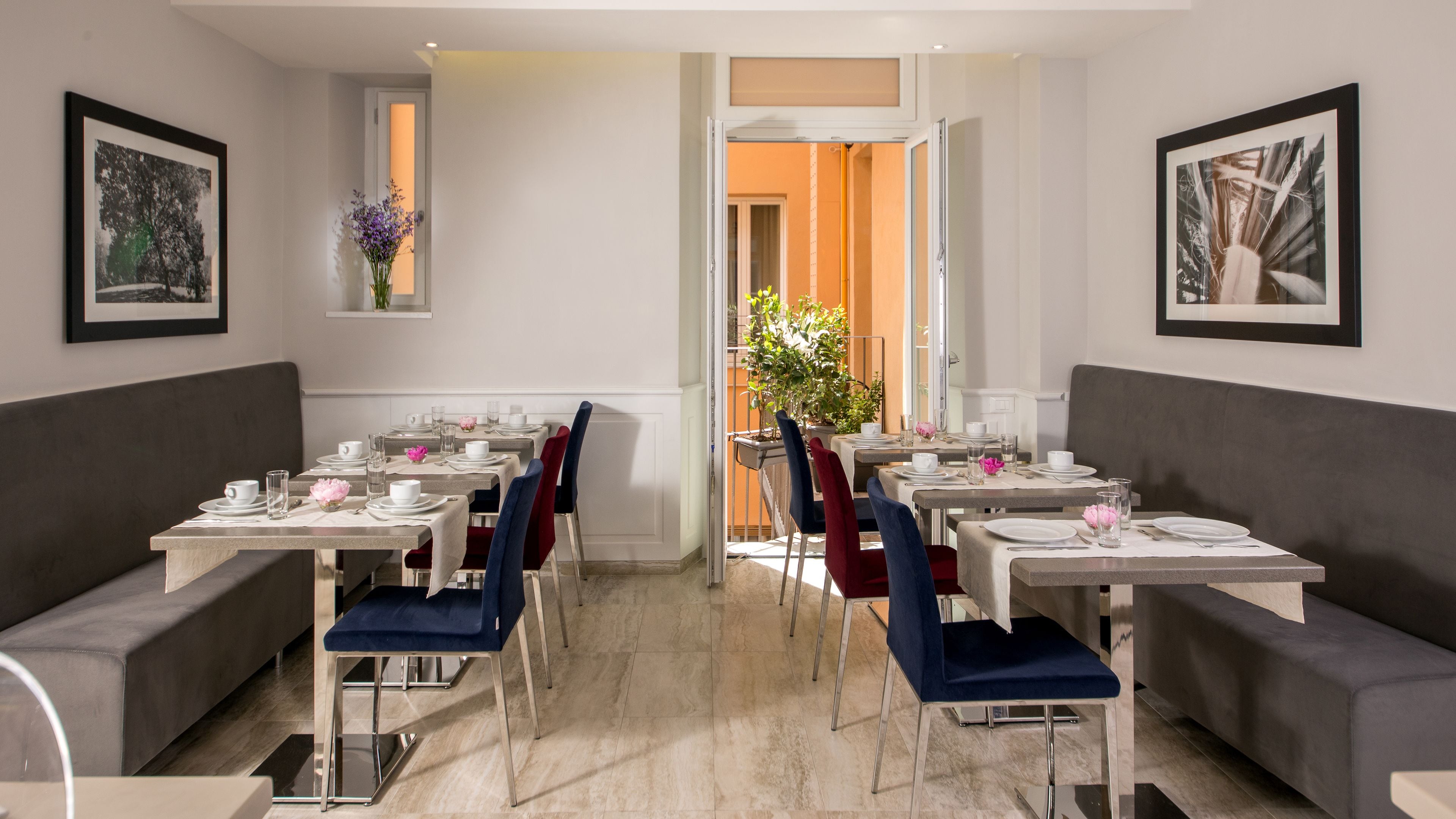Un comedor con mesas dispuestas para cuatro, cada una con platos blancos, vasos y cubiertos. Hay dos sofás y una planta en maceta en las paredes.