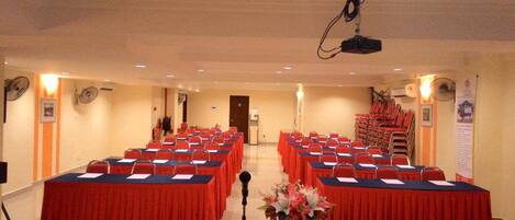 Un salón de conferencias con filas de mesas y sillas, un atril y un proyector.