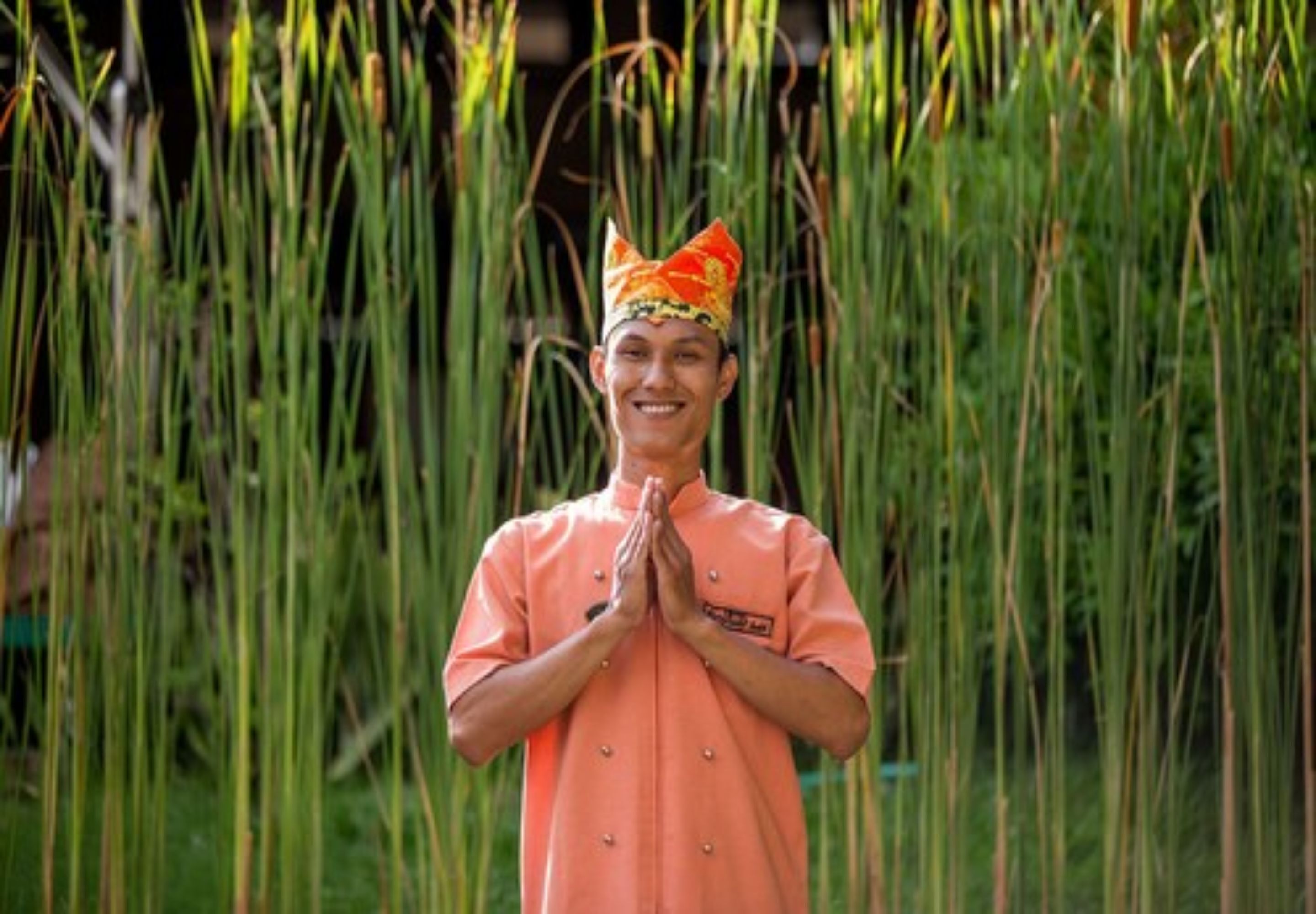 A person in a uniform standing with hands together in front of tall green plants.