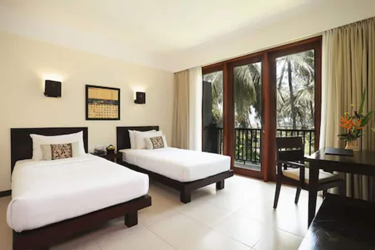 A hotel room with two beds, a dining table, and a balcony with a view of palm trees.