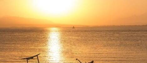 A sunset over a calm sea with boats and people on the shore.