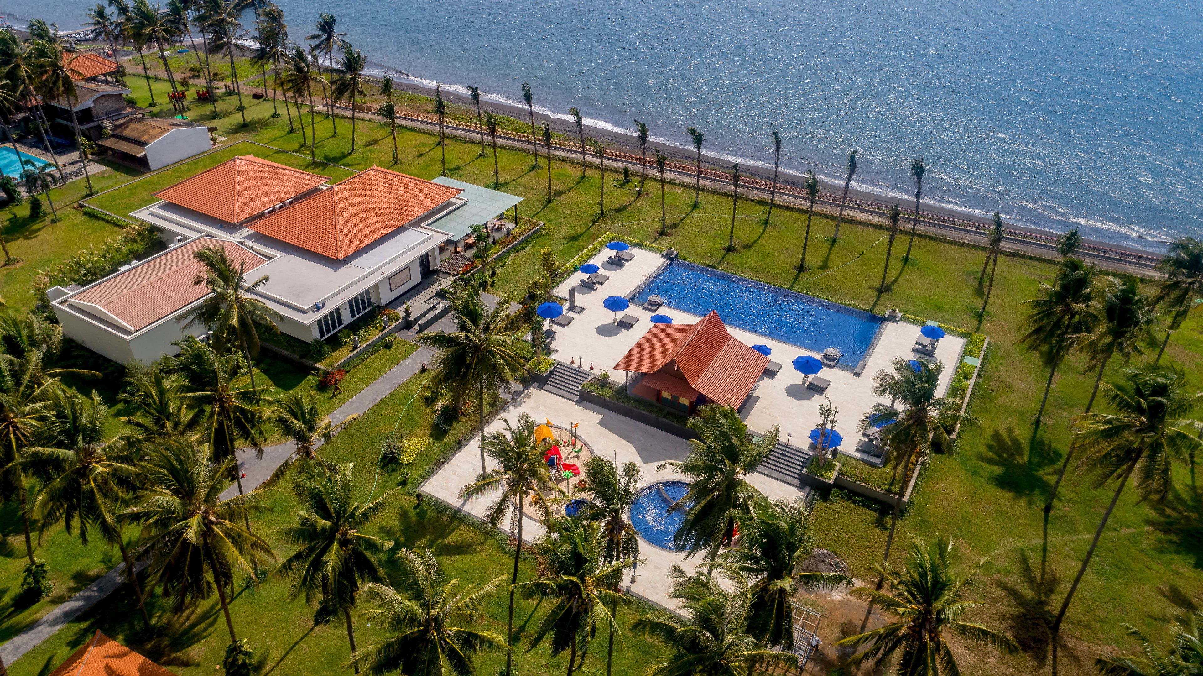 An aerial view of a resort with a swimming pool, palm trees, and a beach.