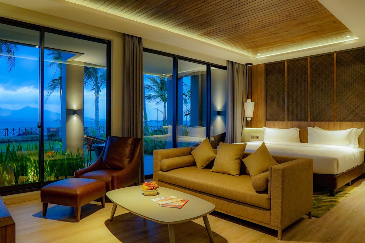 A modern hotel room with a large bed, a sofa, a coffee table, and a view of palm trees and mountains.