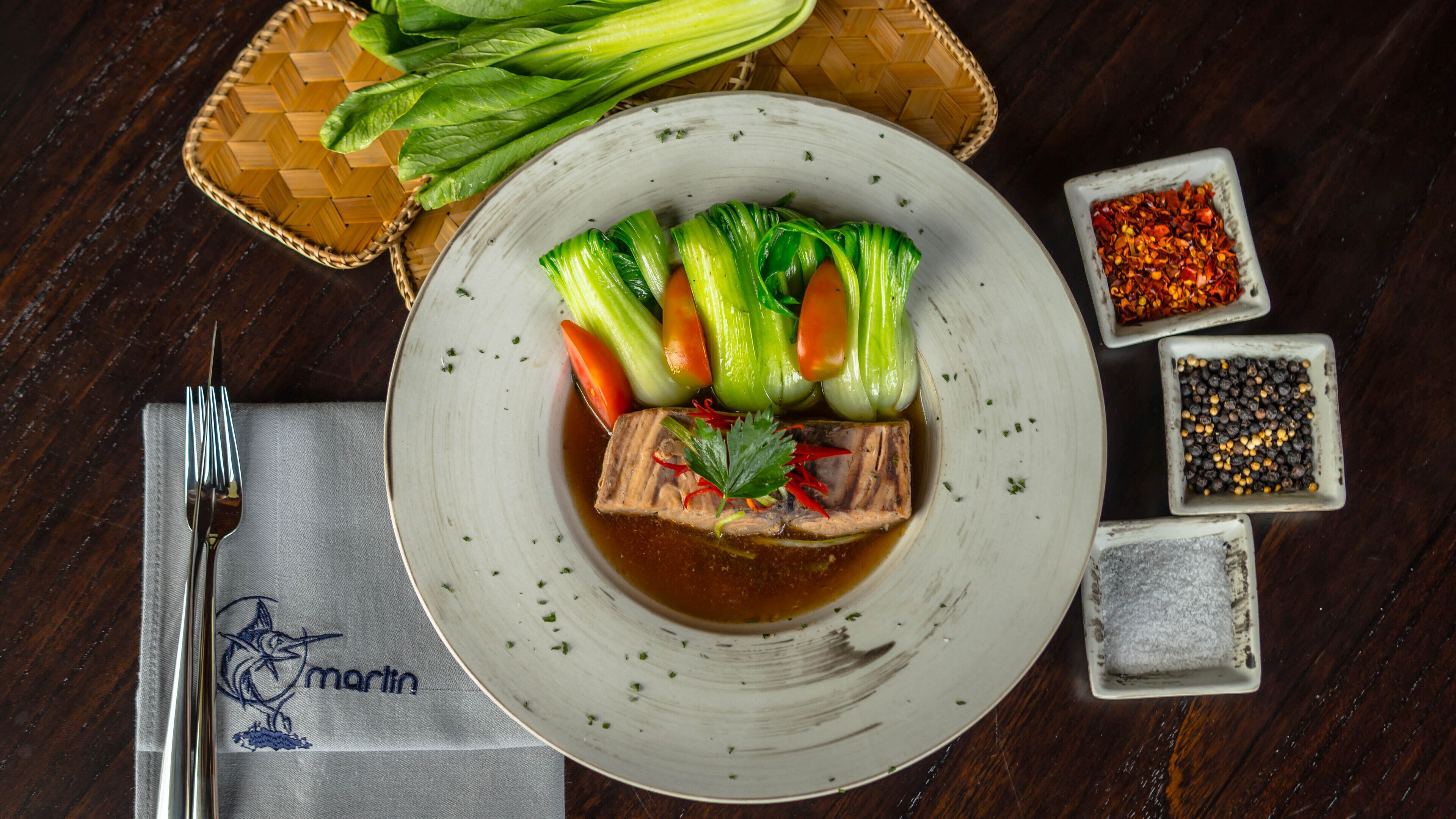 Un plato de comida con verduras y carne, acompañado de un tenedor y una servilleta sobre una mesa de madera.