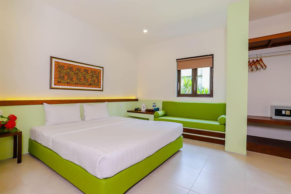 A hotel room with a large bed, a green sofa, a small table with a coffee maker, a window with a view of greenery, and a wall-mounted air conditioning unit.