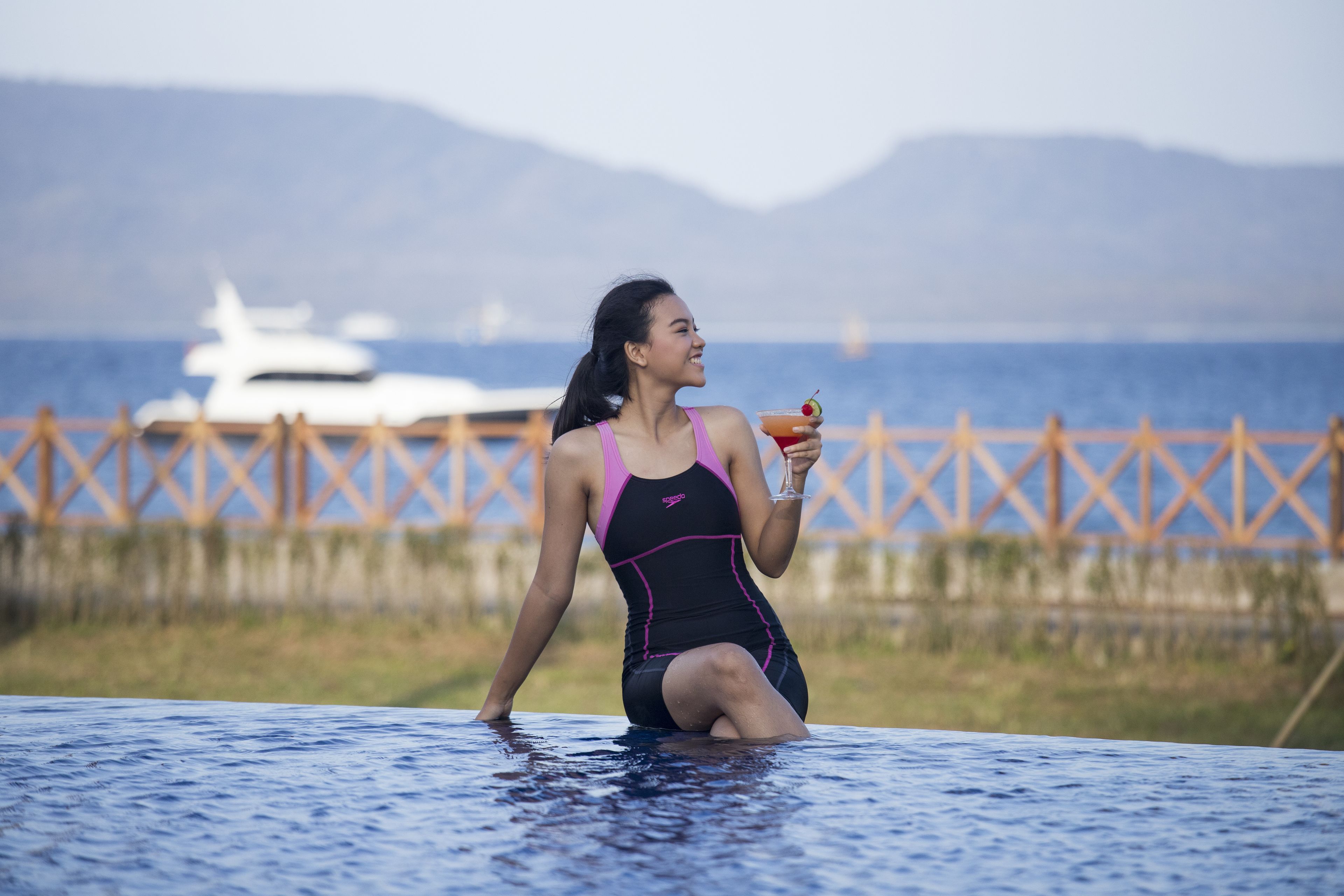 A person in a black and pink swimsuit sitting in a pool with a drink, with a yacht and mountains in the background.