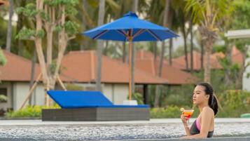 A person in a swimming pool with a blue umbrella and a drink, surrounded by palm trees and a building with a tiled roof.
