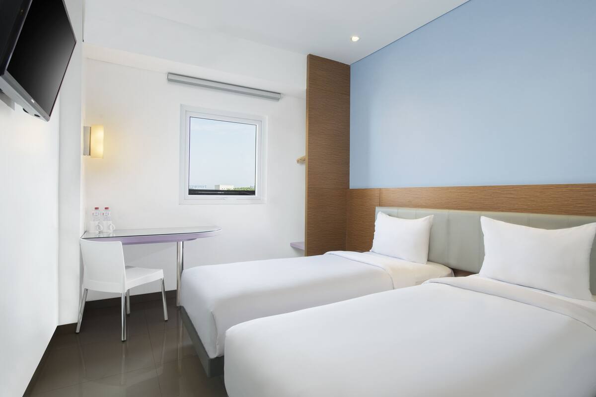Quarto de hotel com duas camas, uma mesa, uma cadeira, uma TV e janela com vista.