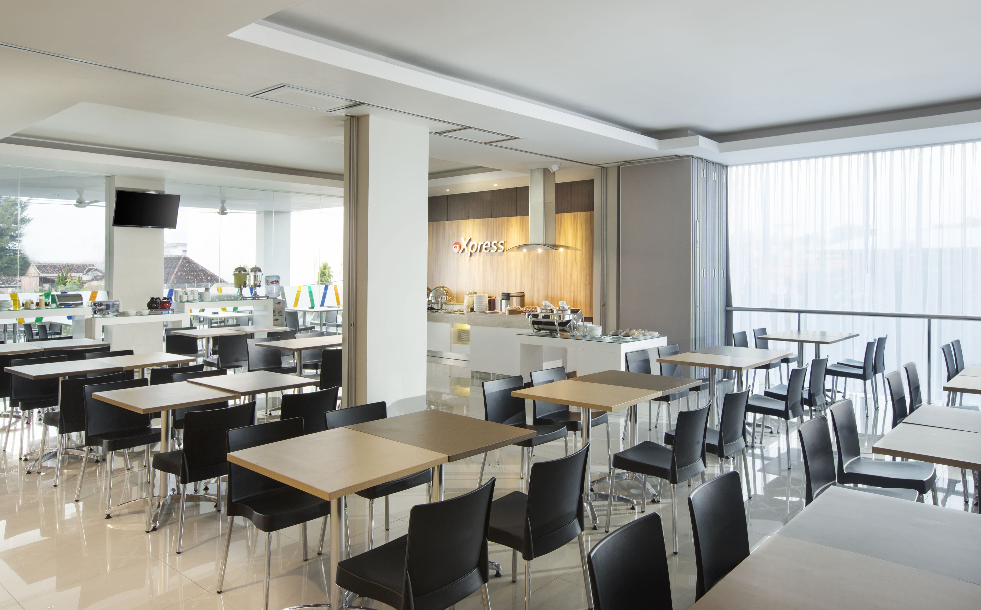 Un espace repas moderne avec plusieurs tables et chaises, un coin cuisine avec une enseigne « Xpress », et de grandes fenêtres offrant une vue sur l’extérieur.